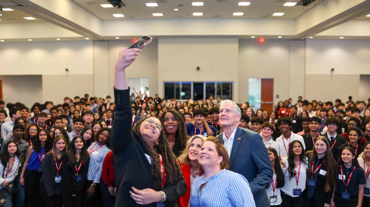 A young woman holds up a smartphone to take a group selfie with adult leaders and a large crowd of smiling high school students in a conference hall during Plano Mayor's Summer Internship Program Prep Day